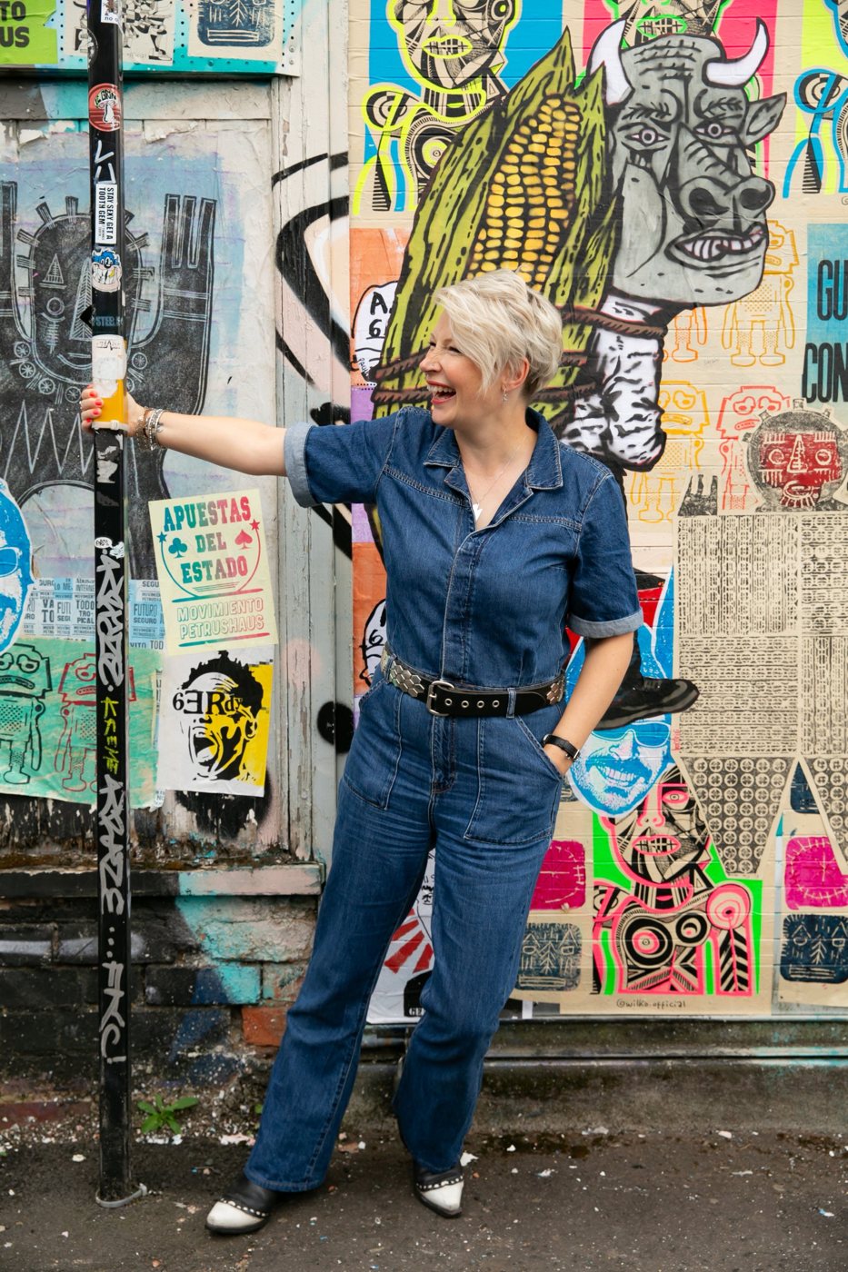 Zoe laughing and swinging from a lamppost in front of a wall of colourful street-art posters