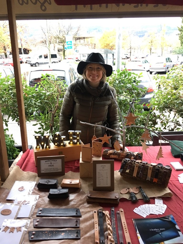 Zoe in a black hat behind her Mischief and Hide market stall, with light-up M and H letters and handmade leather cuffs, wallets and keyrings