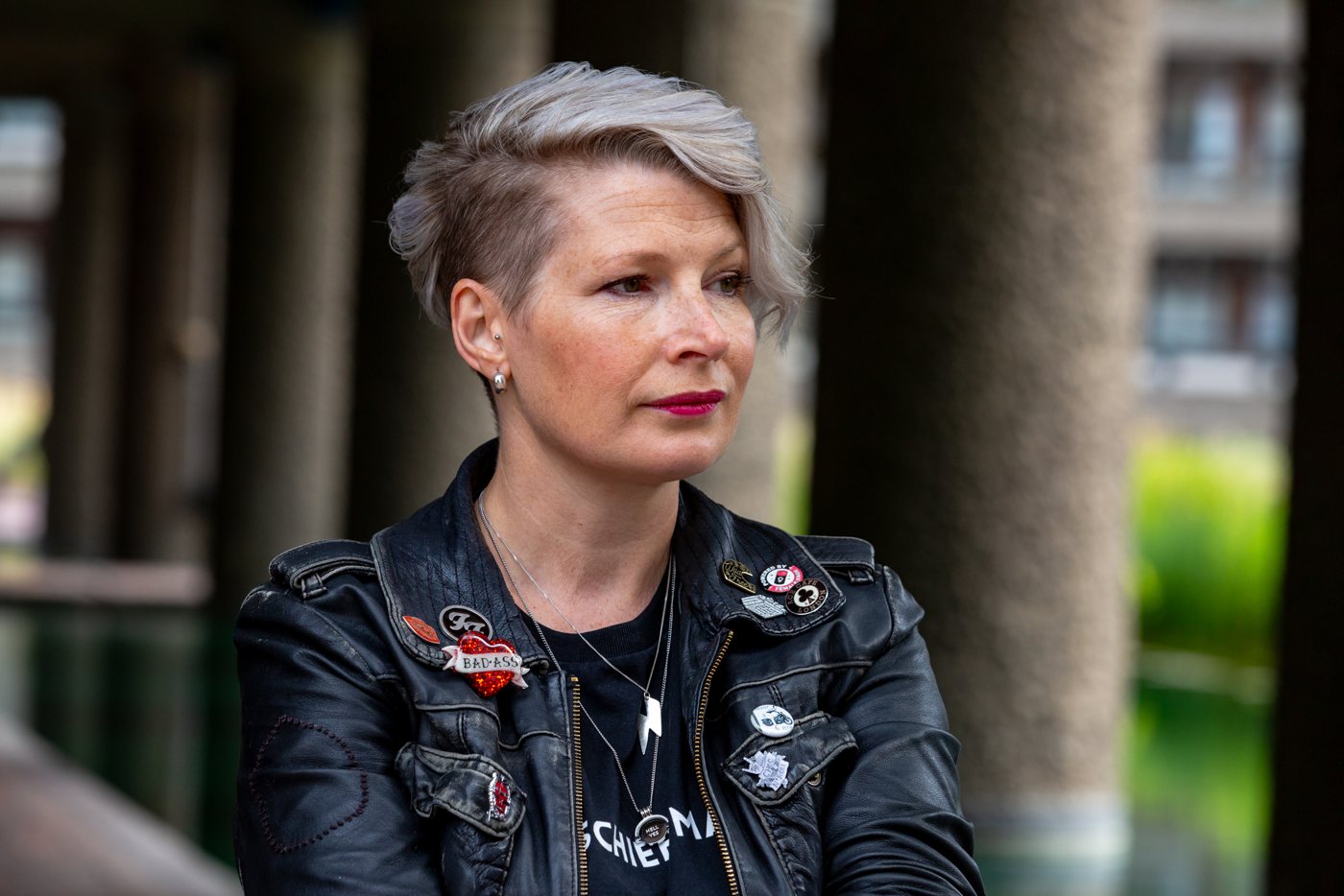 Zoe looking thoughtfully off camera, in a badge-covered leather jacket among concrete columns