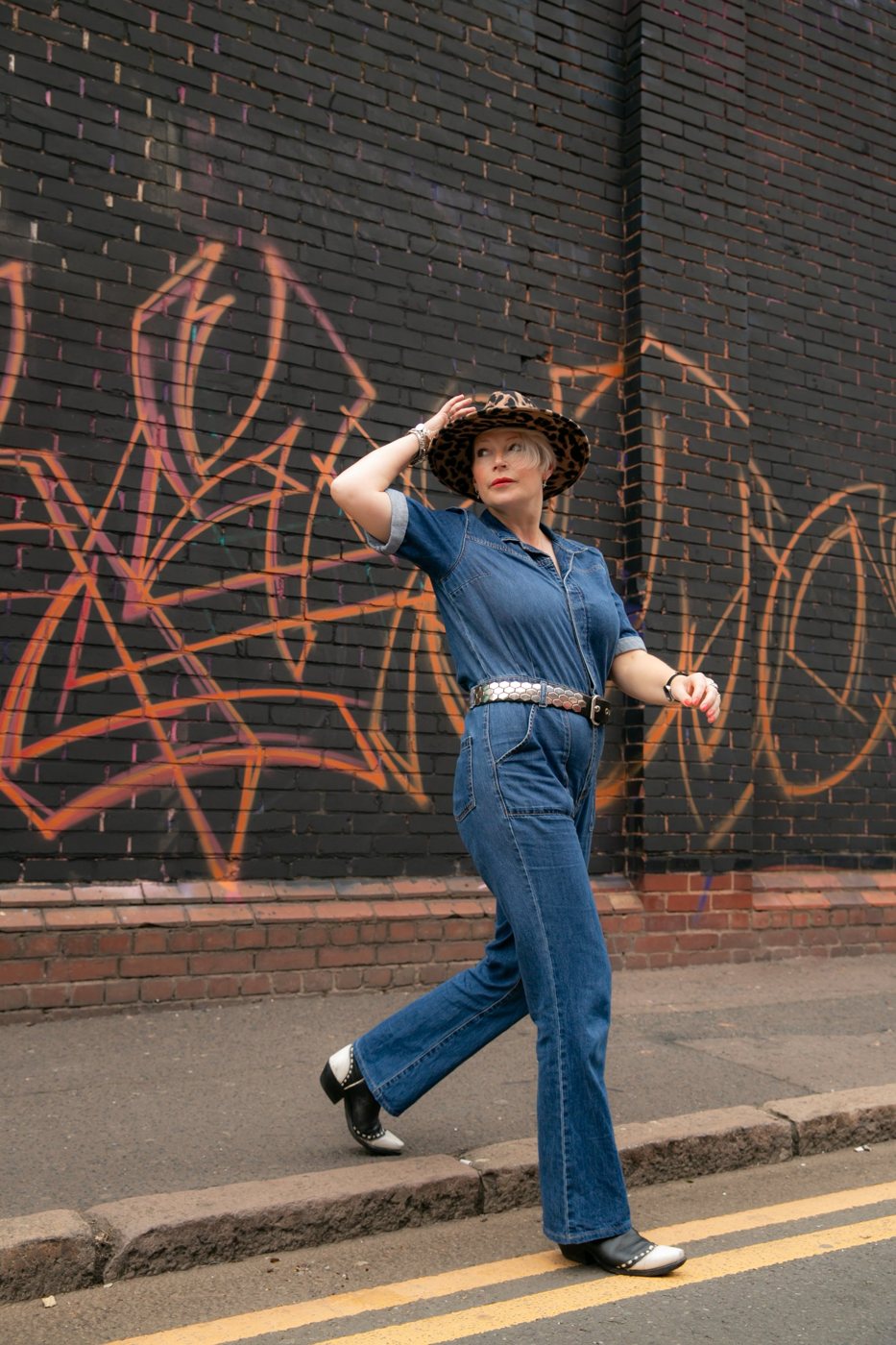 Zoe striding down a street in a denim jumpsuit and leopard print hat, past a graffiti wall